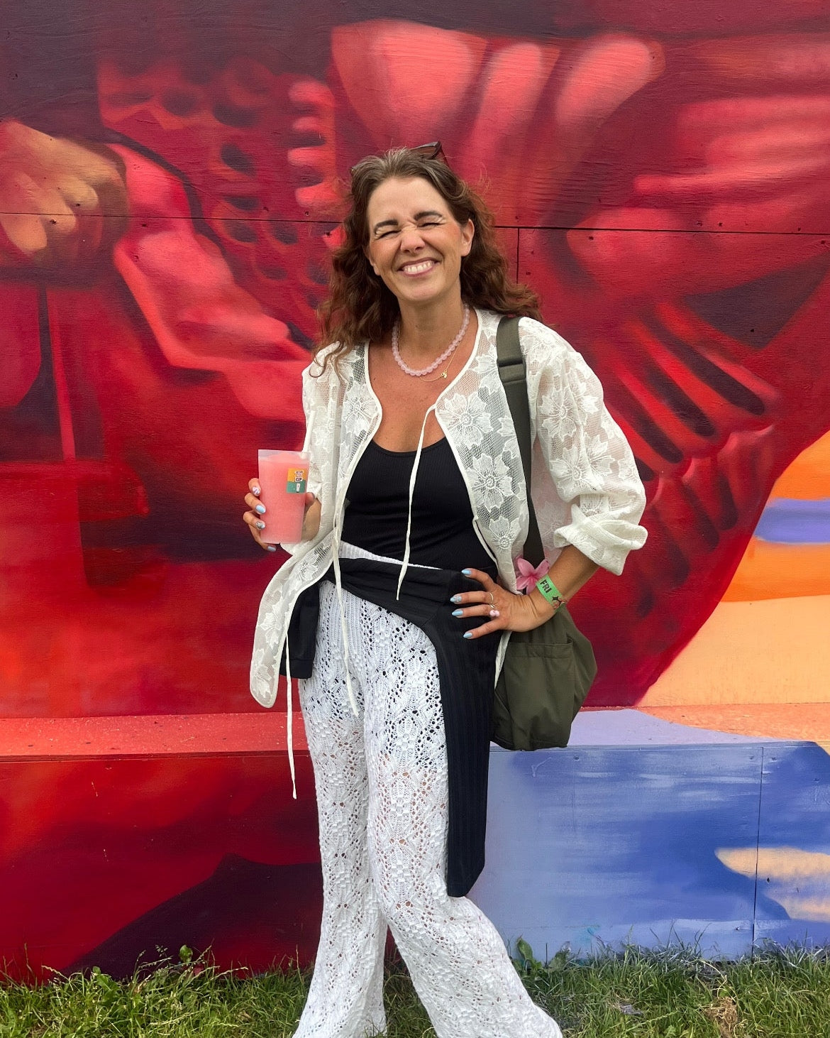 A woman stands outdoors in front of a colorful mural, wearing a black top, white lace wide-leg pants, and a white lace jacket, holding a pink drink.
