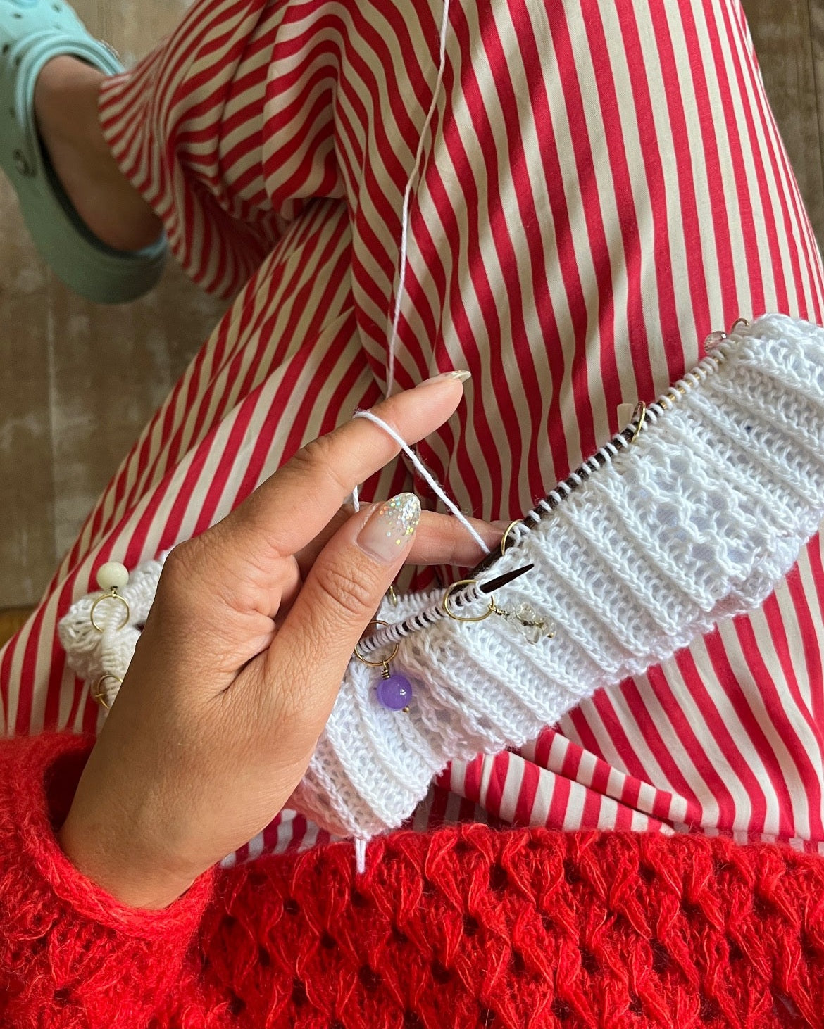 A close-up of a person knitting with white yarn and wooden knitting needles, wearing a red knit sweater, red-and-white striped pants, and a mint green shoe, against a wooden floor background.
