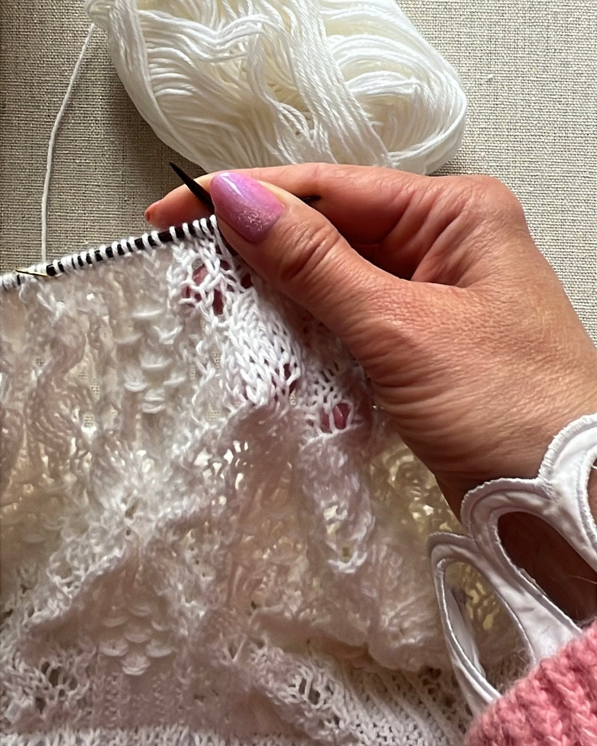A close-up of a hand knitting with white yarn and wooden needles, with a ball of white yarn resting on a beige fabric background.