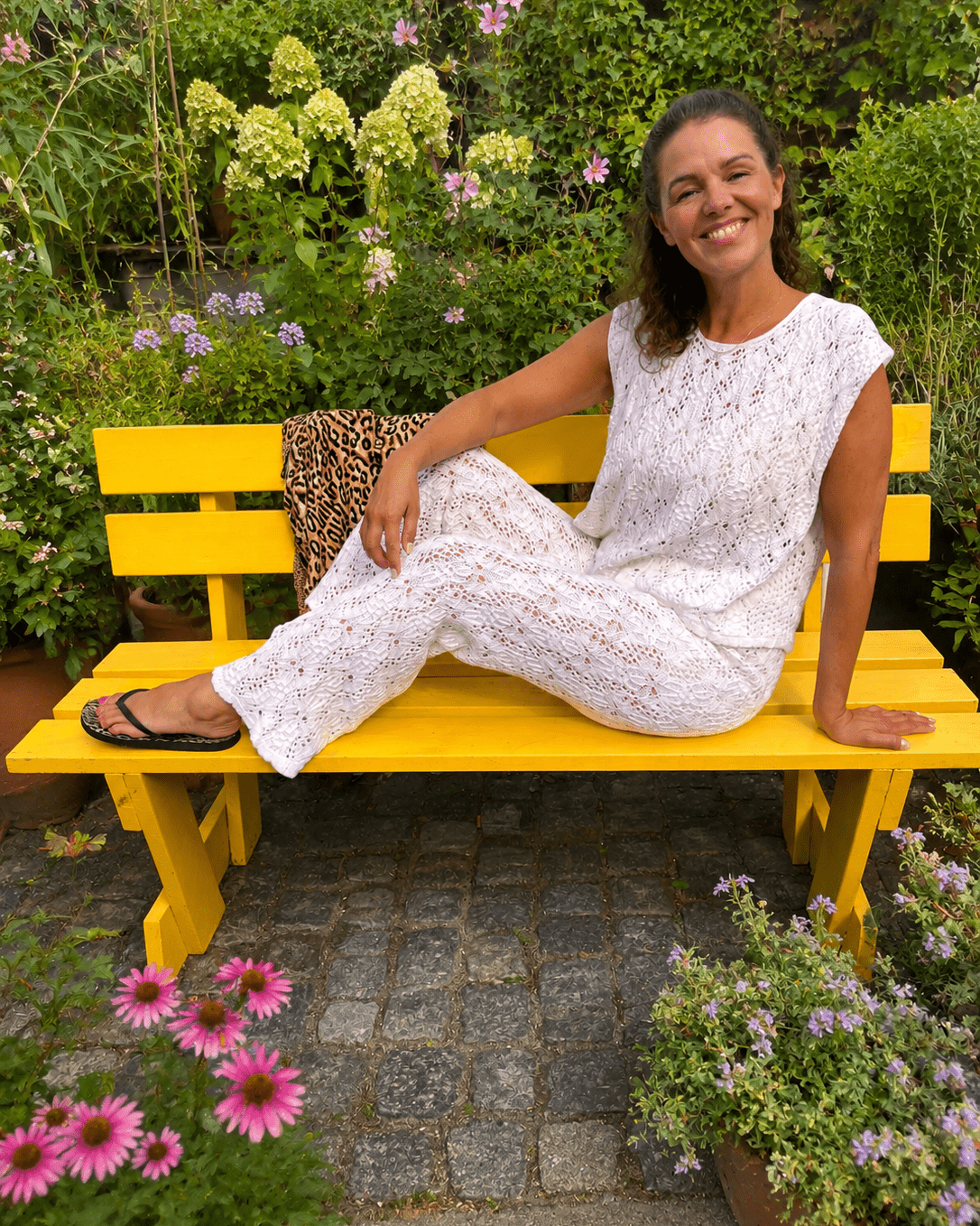 A woman wearing a white knit two-piece set sits on a bright yellow garden bench. The set consists of a short-sleeve crewneck top and matching wide-leg pants.