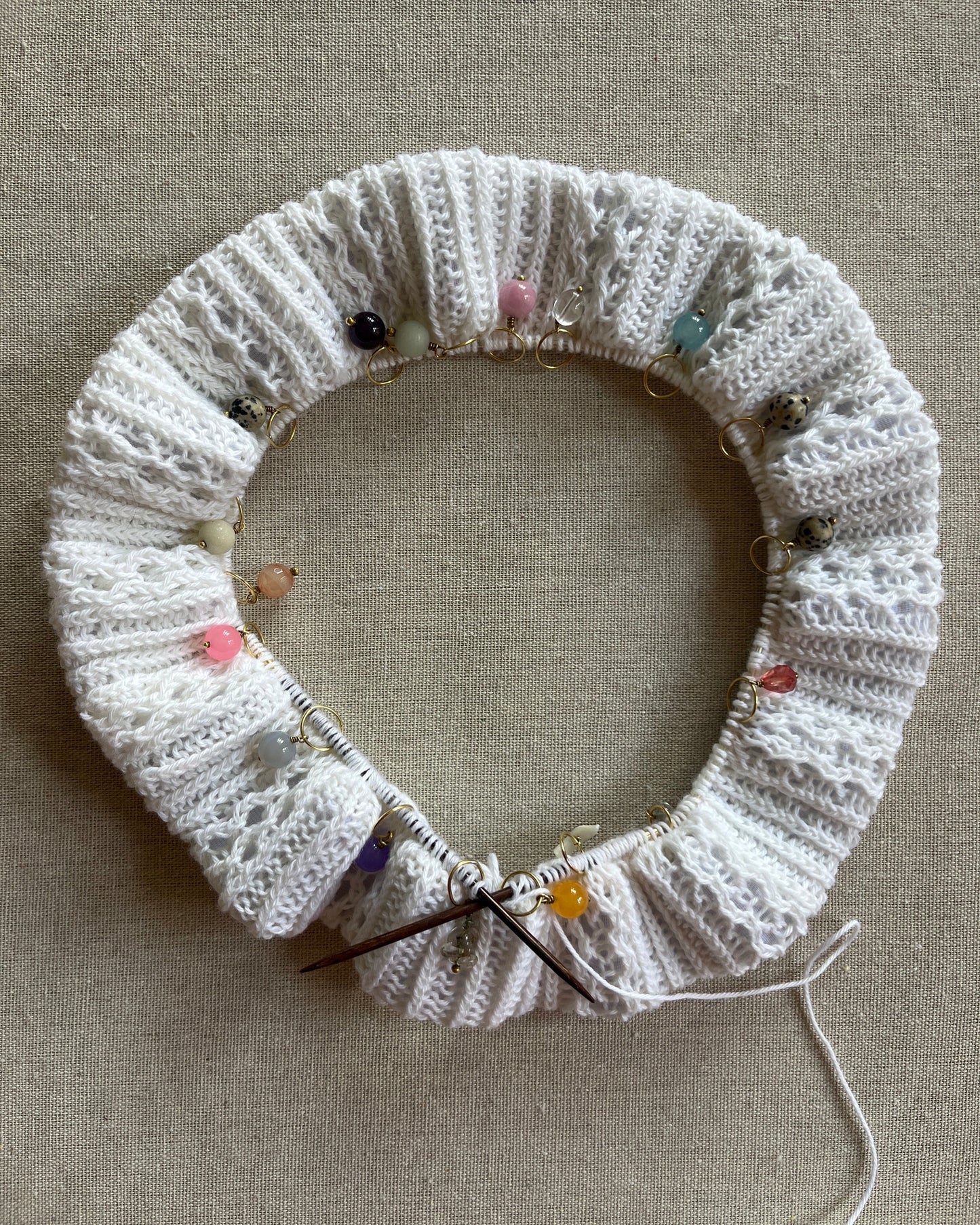 The waistband of the White Nights Pants in the making. On the needle with small colorful stitch markers and a wooden needle, laid flat on a beige linen background