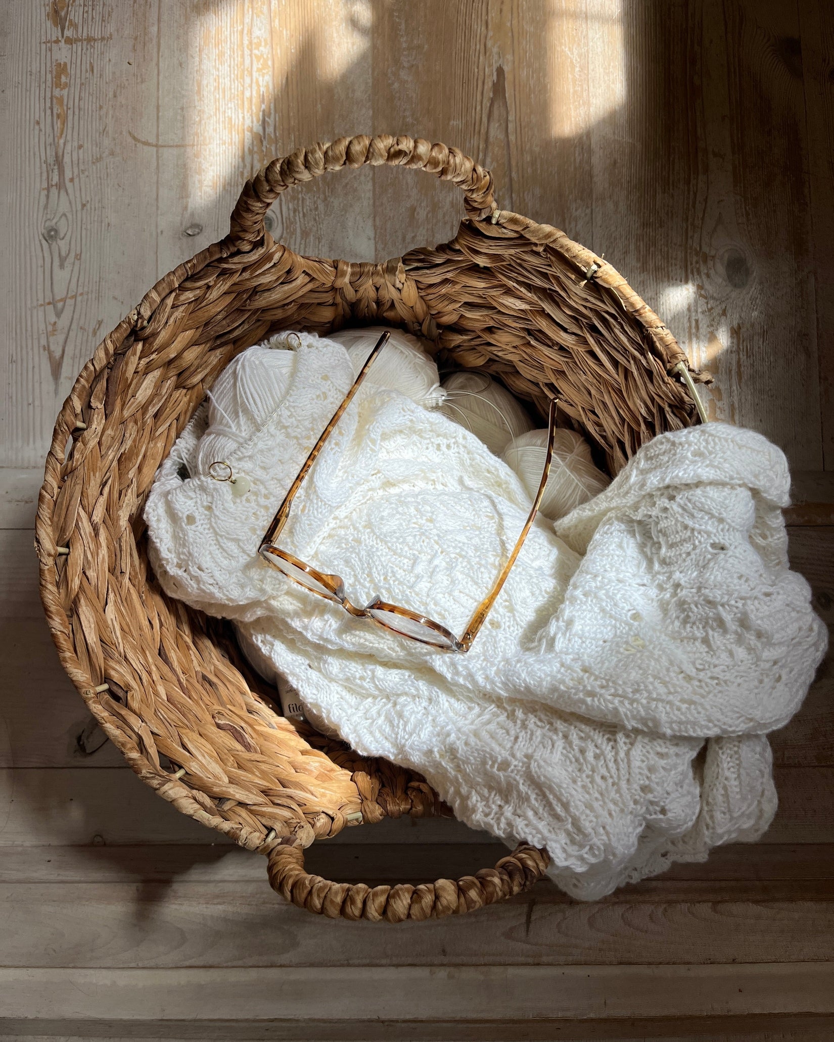 A round woven wicker basket filled with soft white textured knit fabric, with a pair of tortoiseshell eyeglasses resting inside, set against a rustic light wood plank background with dappled sunlight.