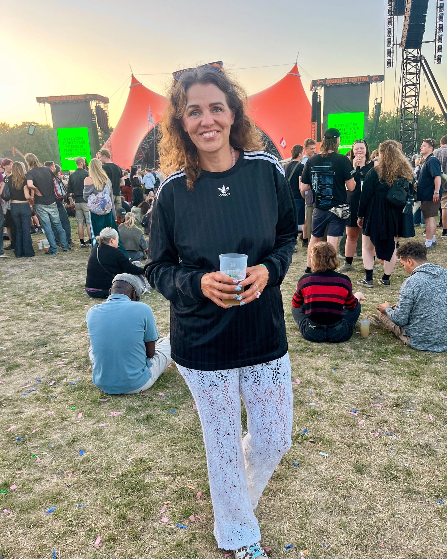 A woman stands on a grassy outdoor festival field at sunset, wearing a black Adidas long-sleeve top with white stripes and white patterned pants, holding a clear plastic cup. An orange stage backdrop and other people are visible in the background.