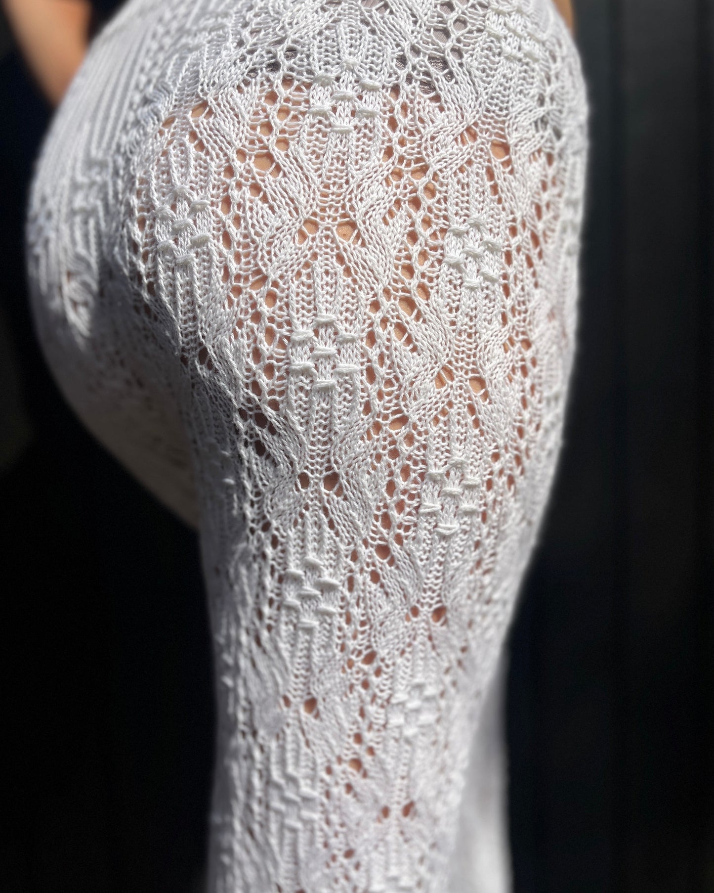 Close-up of a white lace and smock knit garment with an intricate openwork pattern, shown on a person against a dark background.