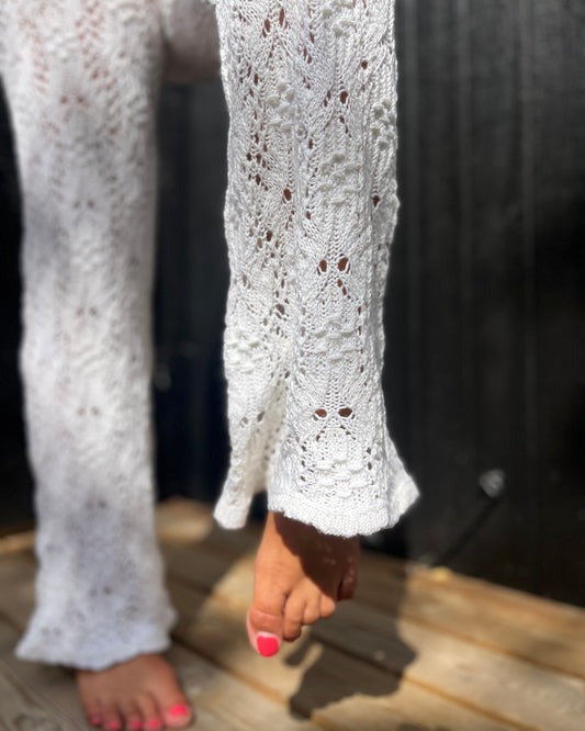 A close-up of a person wearing white open-knit lace and smock pants, standing barefoot on a wooden deck with a dark wooden door in the background.
