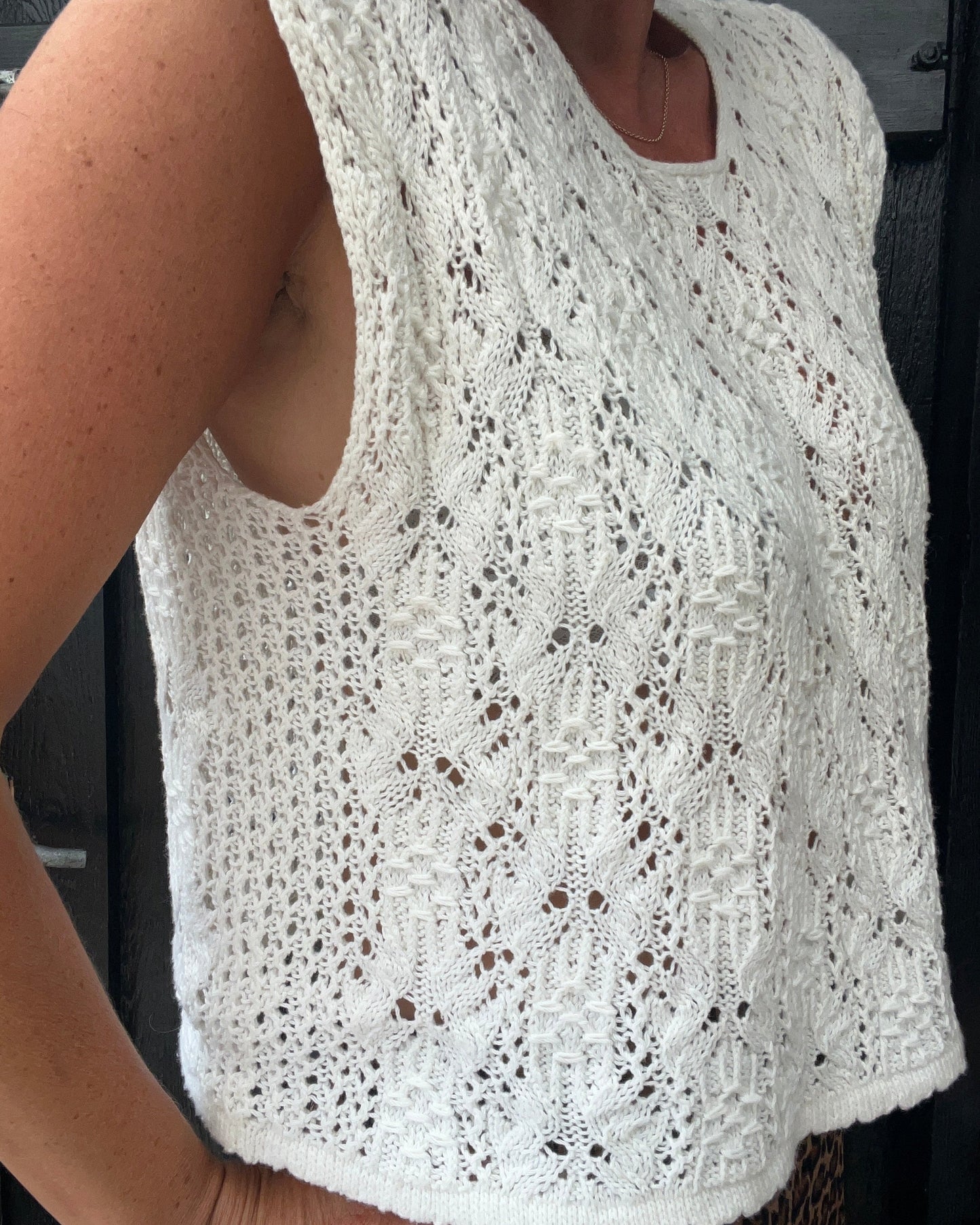 A close-up of a person wearing a sleeveless white lace and smock knit top with an intricate openwork lace pattern.