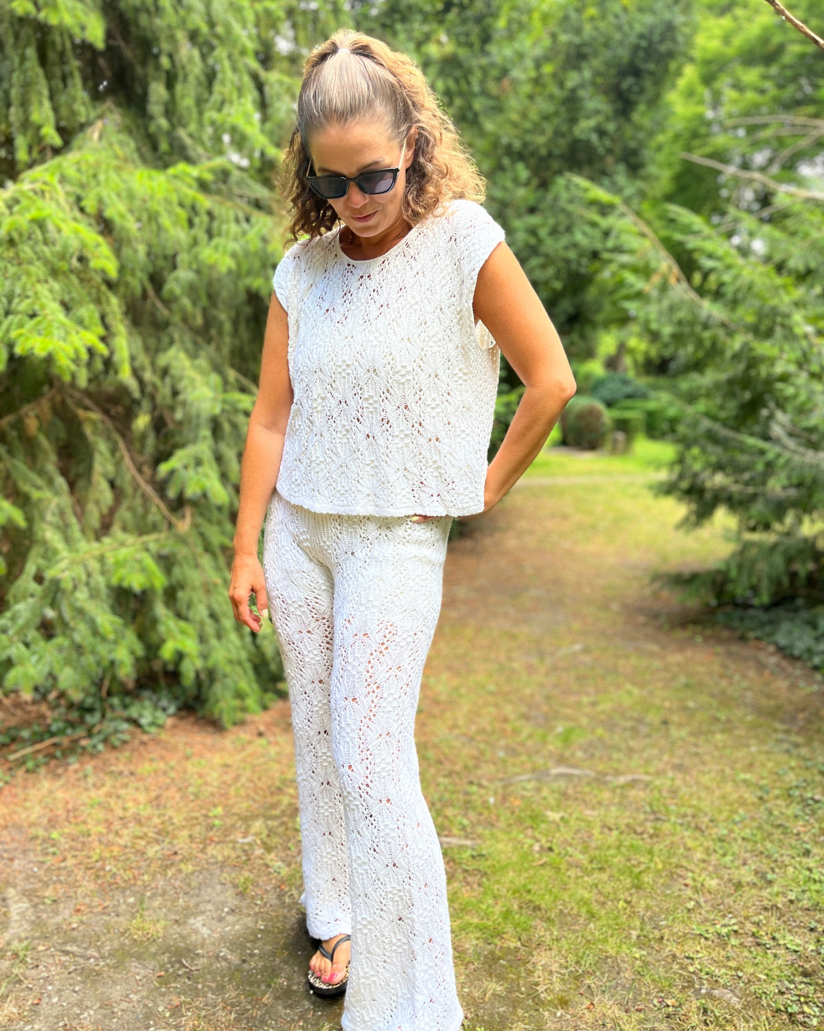 A woman wearing a white lace and smock knit two-piece set consisting of a short-sleeve top and matching wide-leg pants, standing on a grassy path surrounded by green trees.
