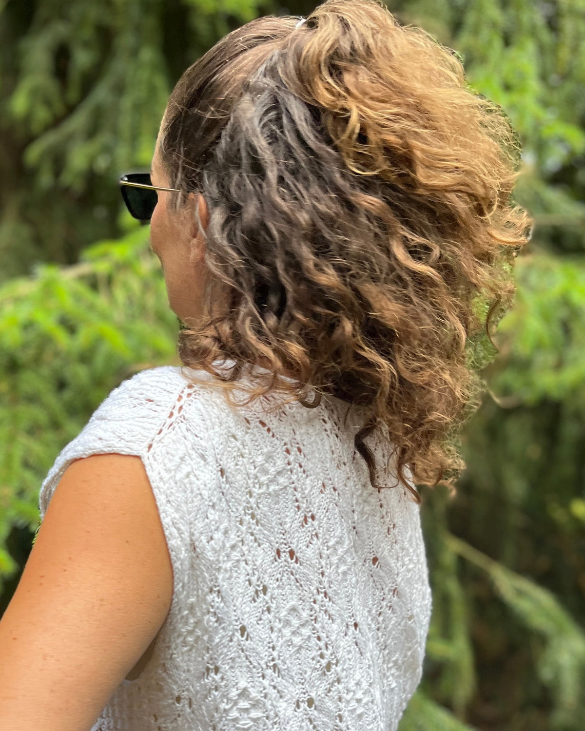 A woman shown from the back wearing a white lace sleeveless top, with curly brown and blonde hair and sunglasses, set against a blurred green forest background.