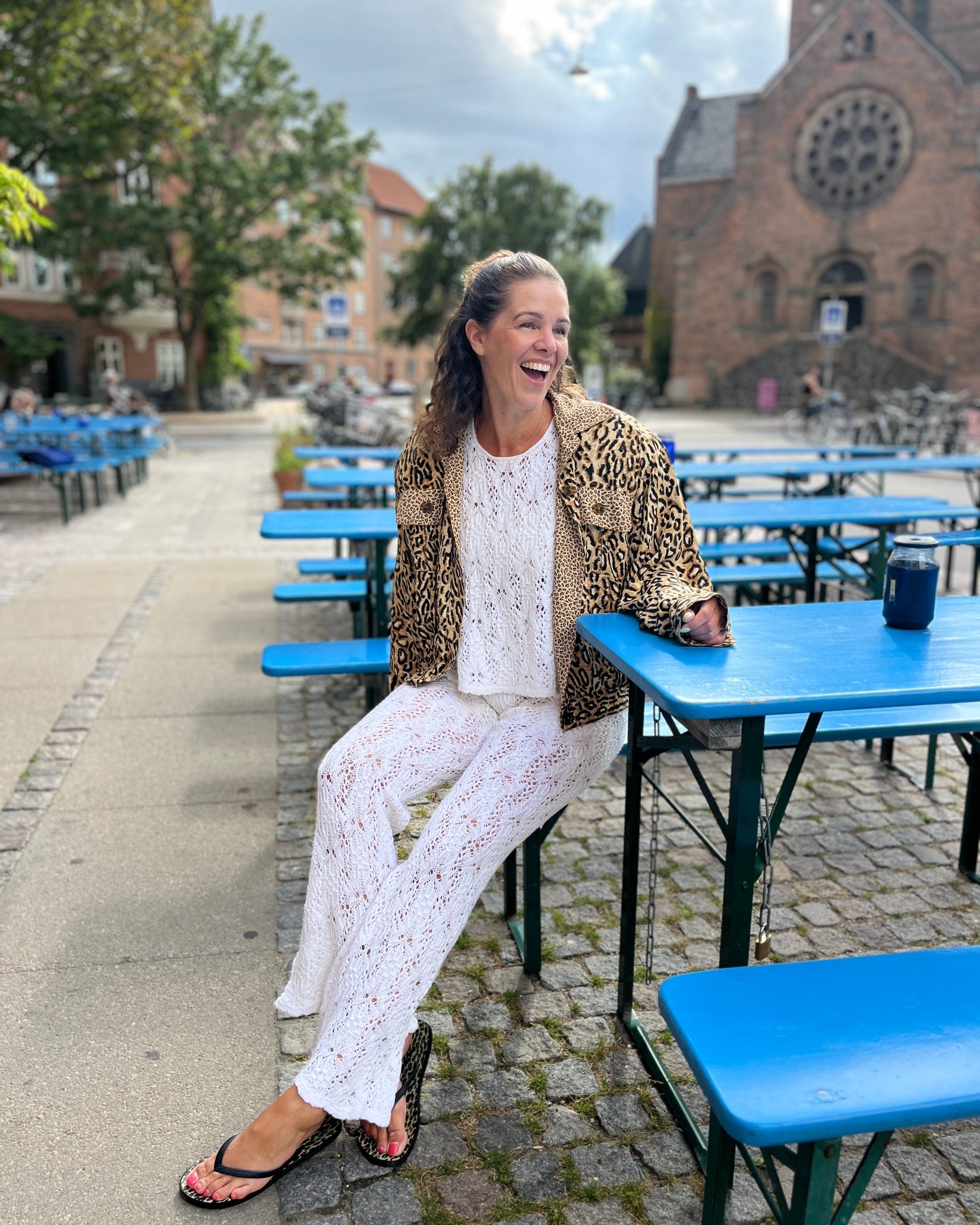 A woman laughing while sitting at a blue outdoor table, wearing a leopard-print jacket over a white lace top and white lace wide-leg pants, with a cobblestone street, church, and more tables in the background.