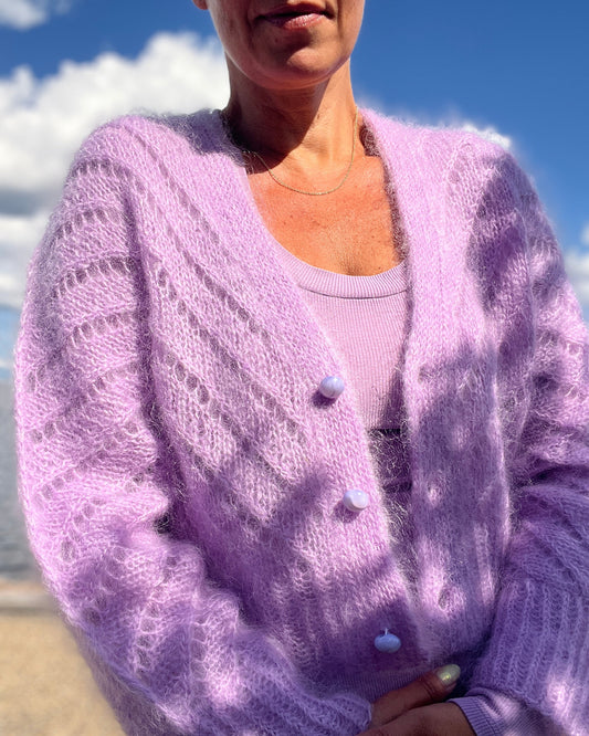 A woman wearing a fuzzy lavender knit cardigan with textured openwork patterns and light-colored buttons, photographed outdoors against a blue sky and sandy beach background.