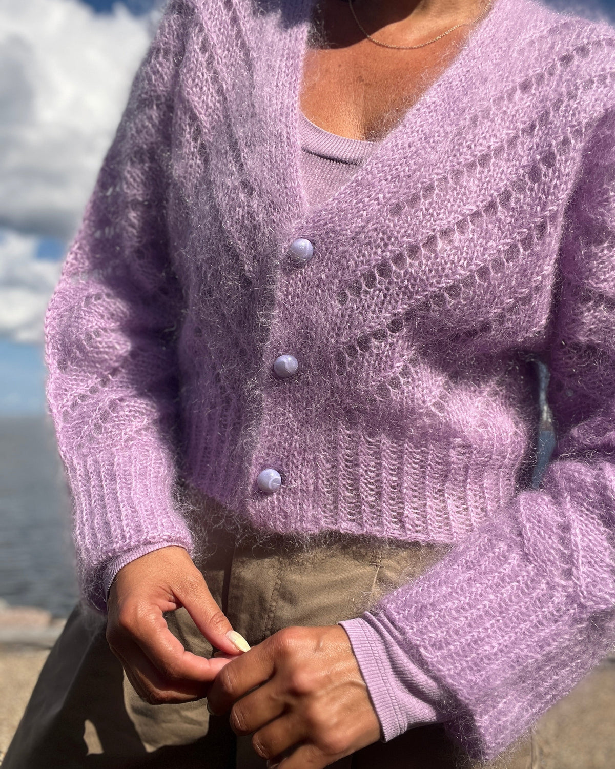 A person outdoors wearing a fuzzy lavender knit cardigan with a V-neck and three front buttons over a matching knit top, paired with khaki pants.