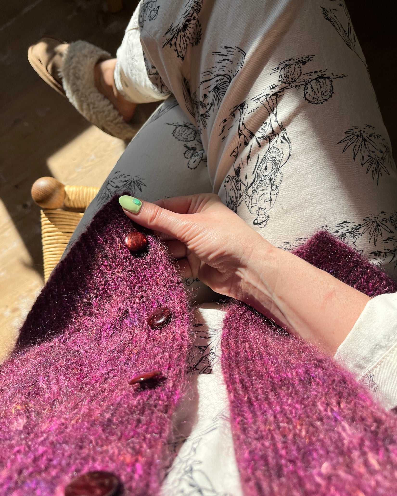Person wearing a patterned shirt and purple cardigan with buttons, sitting on a wooden chair.