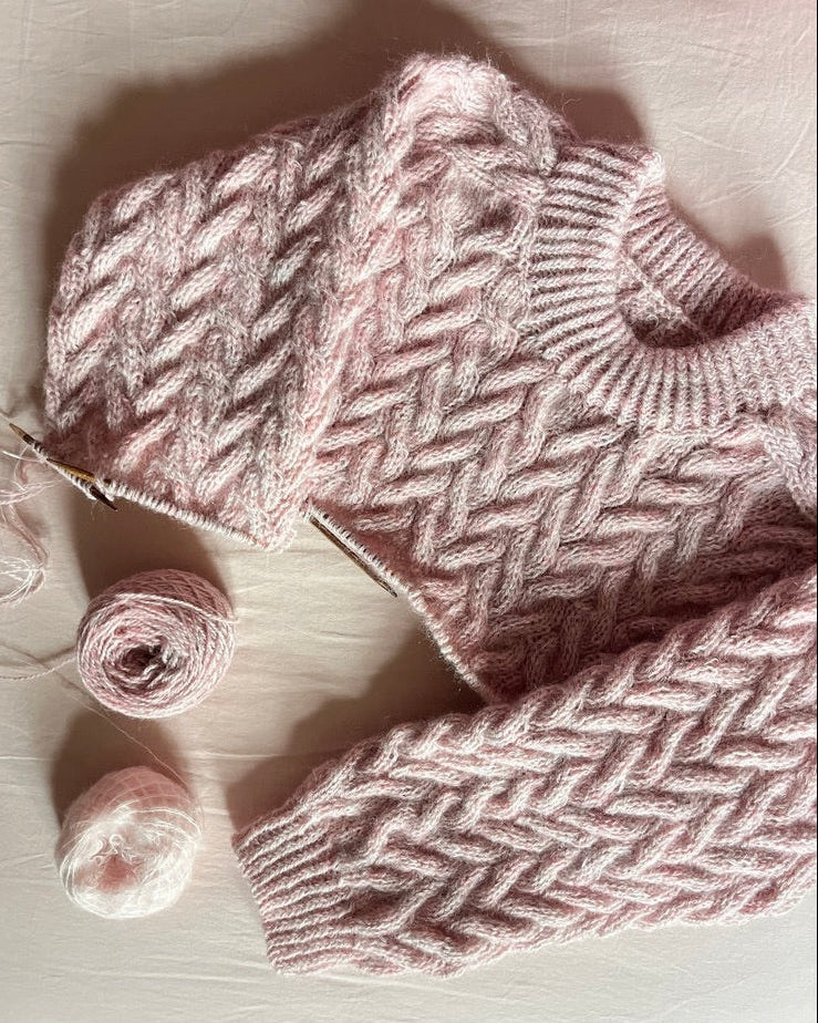 Knitted pink sweater with a herringbone pattern on a light wooden surface, accompanied by two pink yarn balls.