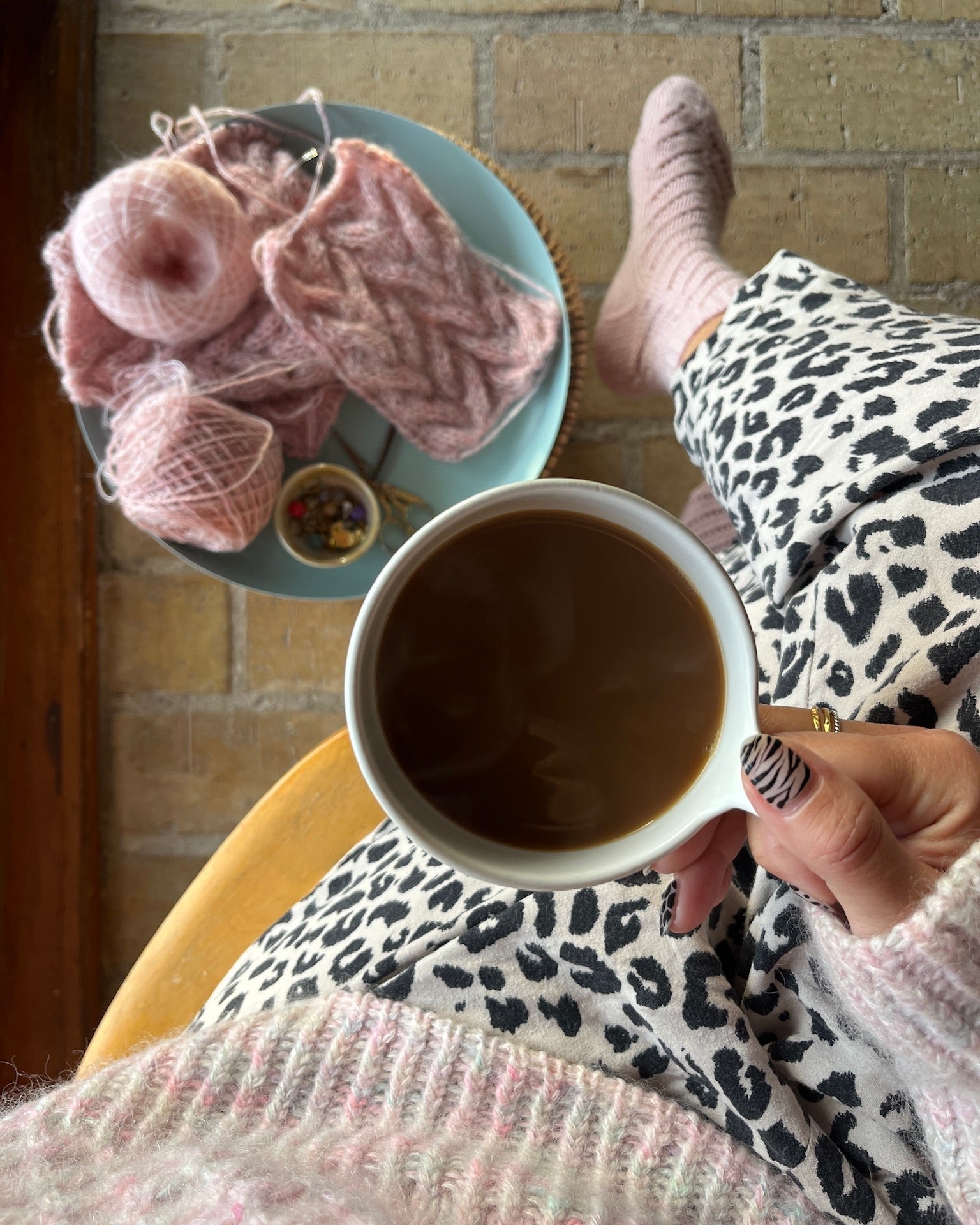Person holding a cup of coffee with pink yarn and knitting needles on a table.