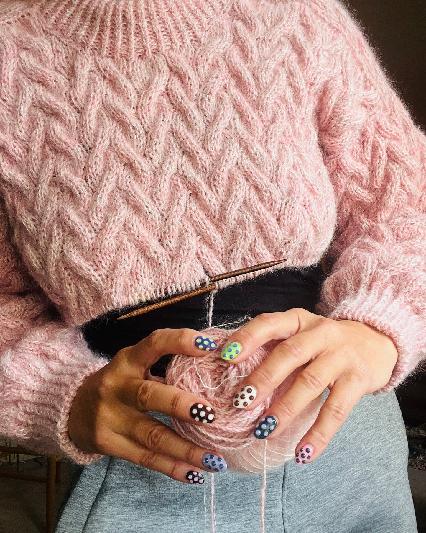 Person wearing a pink knitted sweater holding yarn and knitting needles.