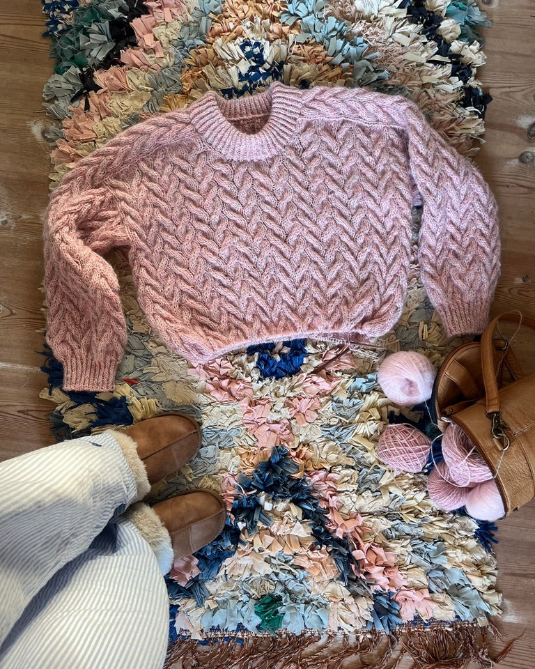 Pink knitted sweater on a colorful textured rug with a handbag and shoes nearby.